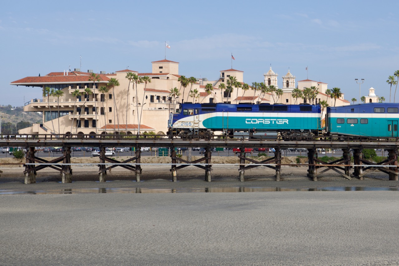 COASTER locomotive 2105 crossing a wooden bridge beside palm trees and a coastal building.